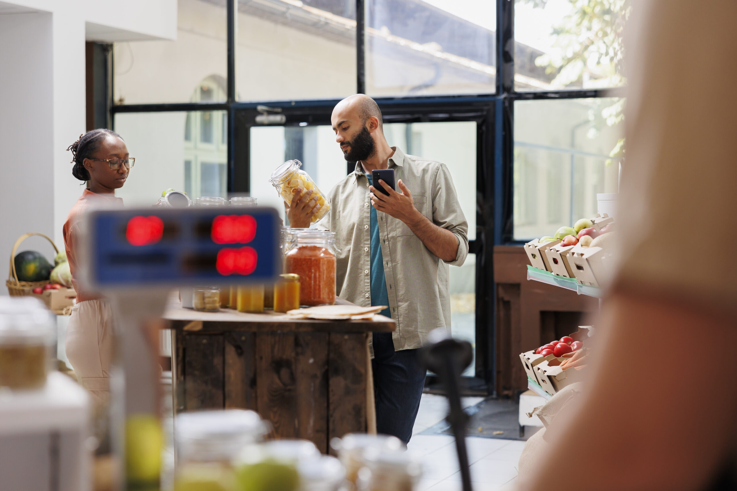Male shopper using mobile device while surrounded by reusable containers and green products. Multiethnic customers utilizing technology, to research local market prices and products.